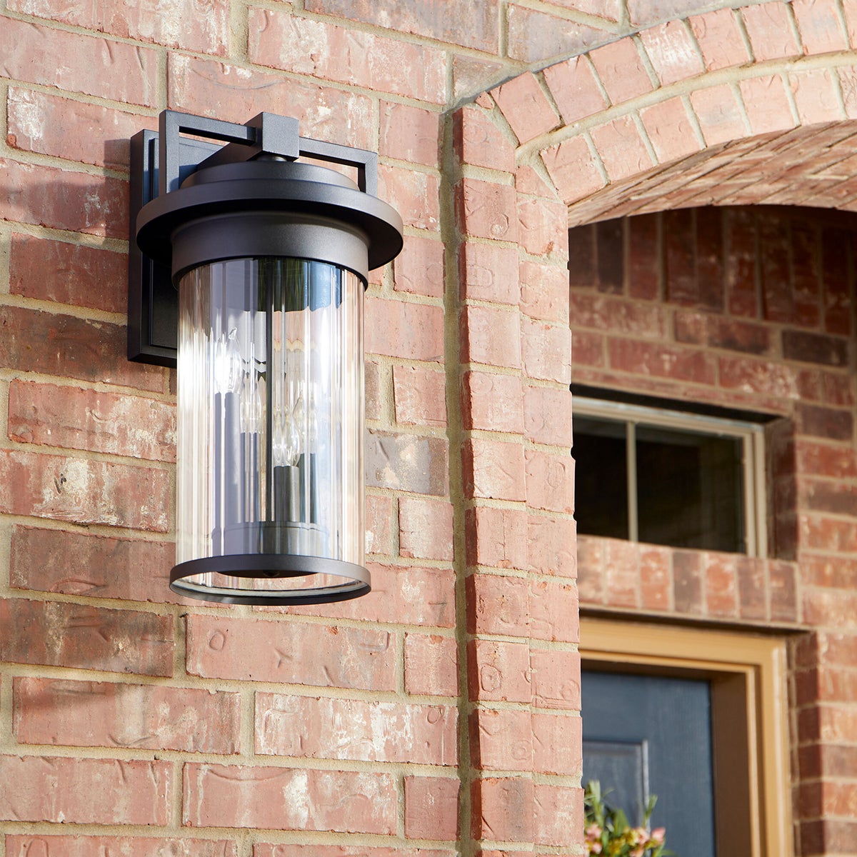 Contemporary outdoor wall light with a cylindrical clear fluted glass shade, crafted from durable metal. Clean lines and a drum-shaped silhouette create a modern touch. Perfect for your outdoor ensemble.