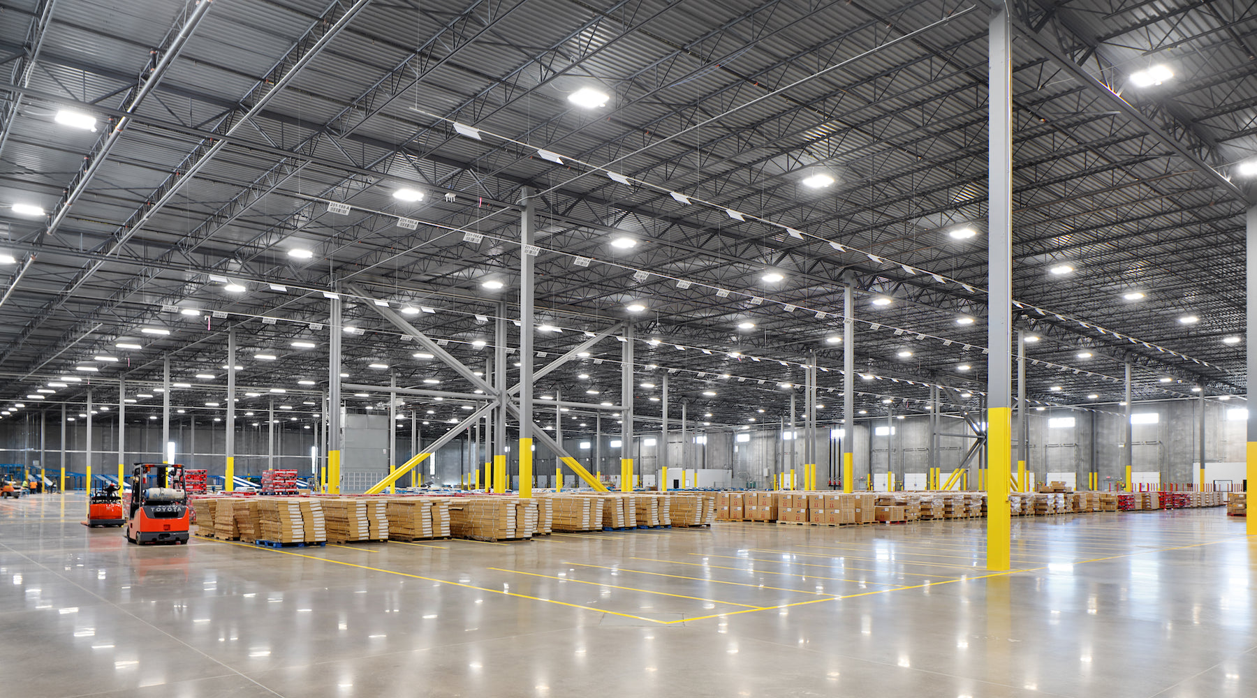 Factory lighting installed in large warehouse with parked forklifts, shipping containers, and crates
