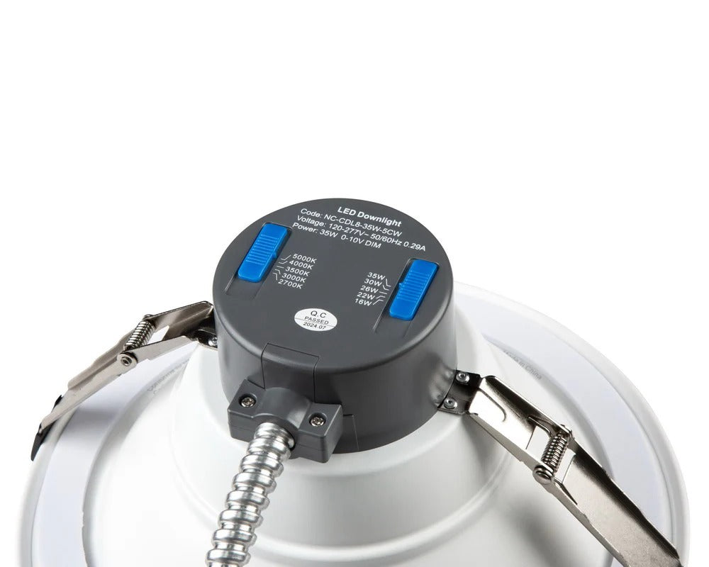 Angled top-down close-up of the 4″ LED commercial downlight showing side conduit entry into the gray top housing, blue selectable wattage/CCT slide switches, and stainless steel mounting clips; 6/8/10/12/16W, 600–1,515 lm, 2700K/3000K/3500K/4000K/5000K, 0–10V dimming, 120–277V.