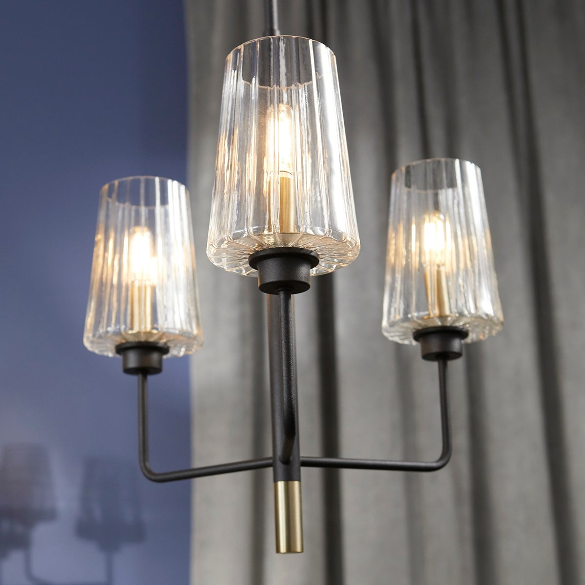 Bathroom chandelier with three clear glass shades, aged brass and noir finish. Linear frame with soft-angular curves. Adjustable chain/stem hung suspension system. Suitable for bathrooms, outdoor kitchens, and covered patios.