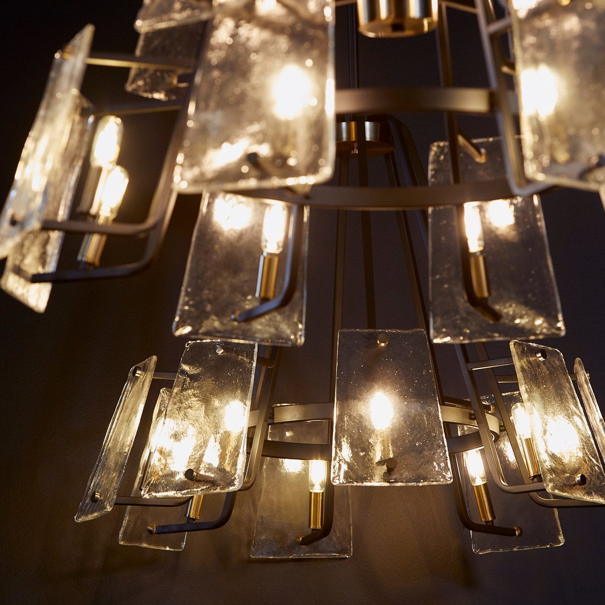 Outdoor chandelier with conical shape, frosted glass trapezoid shades, and aged brass-noir finish. Linear framing and angular accents create a mid-century modern inspired design. Features 12 light sockets orbiting the interior ring. Suitable for covered outdoor spaces or washrooms.