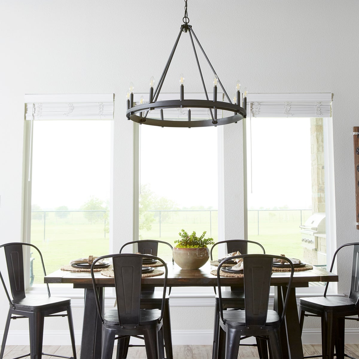 A farmhouse-inspired dining table with chairs and a Wagon Wheel Chandelier, creating an industrial aesthetic. Elevate your space with this customizable fixture featuring a beautiful finish and candle sleeves in aged brass, noir, and satin nickel options. Perfect as a prominent display or supplementary accent.