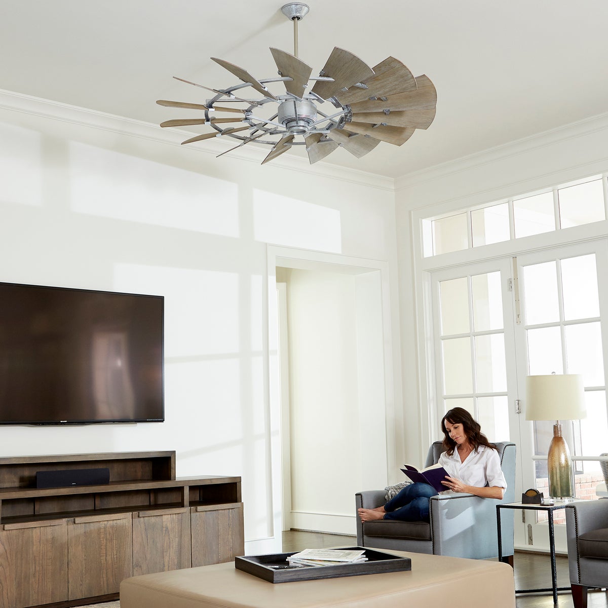 Windmill Ceiling Fan in a rustic living room with a woman sitting in a chair reading a book. Wooden blades, country chic design.
